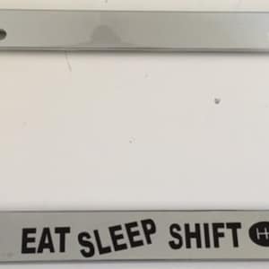 Könnte beinhalten: Chromfarbener Nummernschildrahmen mit dem Text "EAT SLEEP SHIFT" und einem Schaltknauf-Symbol.