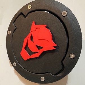 May include: Black gas cap cover with a red stylized animal head design.