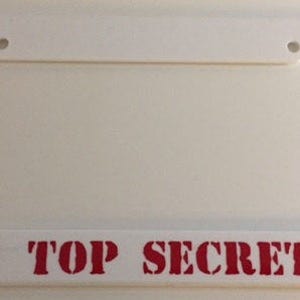May include: White plastic license plate frame with red lettering that says "TOP SECRET".