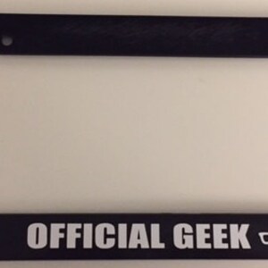 May include: Black plastic license plate frame with white text that reads "OFFICIAL GEEK" and a pair of glasses icon.