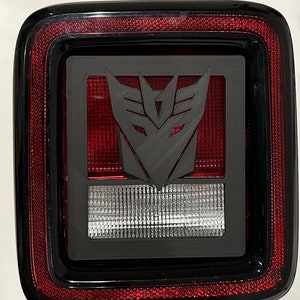 May include: A black plastic automotive taillight with a red lens and a gray 3D printed emblem of the Transformers Autobot symbol.