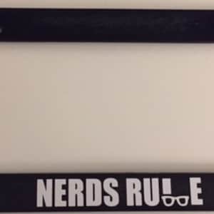 May include: Black plastic license plate frame with white text that reads "NERDS RULE" and a pair of glasses icon.