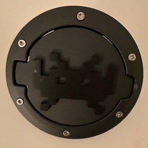 Può includere: Un tappo del serbatoio rotondo nero con un design a forma di astronave argentata. Il tappo del serbatoio ha sei viti argentate attorno al bordo.