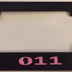 May include: Black license plate frame with a pink silhouette of a head and the number 011.