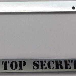 May include: A chrome license plate frame with a white background and black text that reads "TOP SECRET".