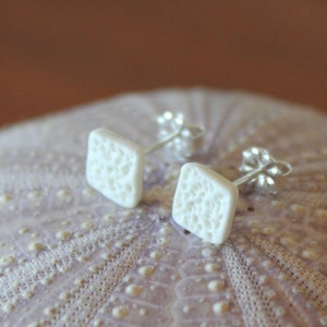 May include: A pair of white, square stud earrings with a textured surface. The earrings are set on silver posts. The earrings are displayed on a light purple, textured surface.