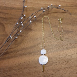 May include: A gold chain necklace with a white ceramic pendant featuring two circles and a long, thin gold bar.
