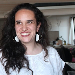 Op de afbeelding: Een vrouw met donker krullend haar lacht, met grote gouden oorringen met witte halve maan accenten. Ze draagt een wit geborduurd topje. De achtergrond is een heldere, binnenomgeving.
