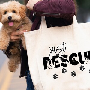 K&ouml;nnte beinhalten: Eine cremefarbene Tragetasche mit dem schwarzen Schriftzug "just RESCUE", Hundesilhouetten und Pfotenabdr&uuml;cken. Ein kleiner, flauschiger, hellbrauner Hund wird vor der Tasche gehalten.
