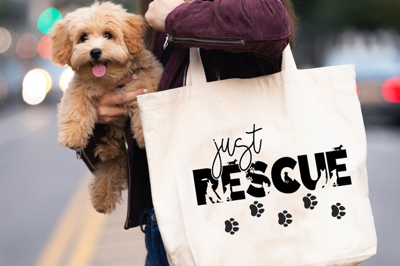 K&ouml;nnte beinhalten: Eine cremefarbene Tragetasche mit dem schwarzen Schriftzug "just RESCUE", Hundesilhouetten und Pfotenabdr&uuml;cken. Ein kleiner, flauschiger, hellbrauner Hund wird vor der Tasche gehalten.