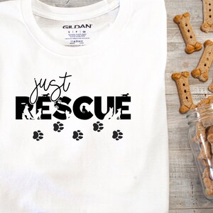 K&ouml;nnte beinhalten: Wei&szlig;es T-Shirt mit dem schwarzen Schriftzug "just RESCUE", Hundesilhouetten und Pfotenabdr&uuml;cken. Knochenf&ouml;rmige Hundekekse liegen neben einem Glasgef&auml;&szlig; auf einer Holzoberfl&auml;che.