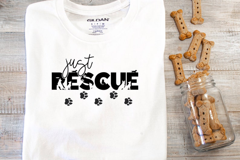 K&ouml;nnte beinhalten: Wei&szlig;es T-Shirt mit dem schwarzen Schriftzug "just RESCUE", Hundesilhouetten und Pfotenabdr&uuml;cken. Knochenf&ouml;rmige Hundekekse liegen neben einem Glasgef&auml;&szlig; auf einer Holzoberfl&auml;che.