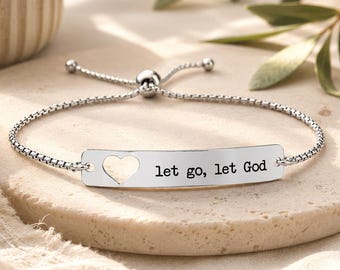May include: A silver-toned adjustable bracelet featuring a rectangular metal bar with a cutout heart shape on the left side and the engraved text "let go, let God" on the right. The bracelet has a delicate box chain and a sliding bead closure.