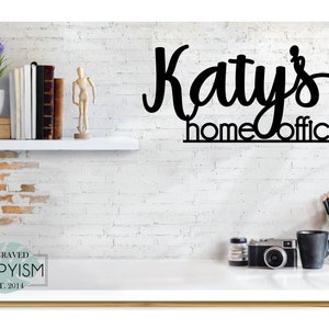 May include: Black metal sign with the text "Katy's home office" on a white brick wall background. The sign is hanging above a white desk with a camera, a mug, and a jar of pencils.