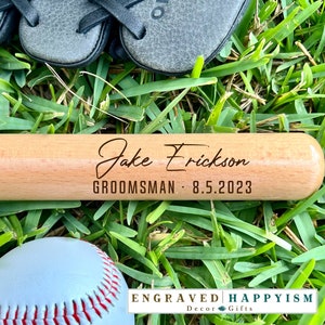 May include: A wooden baseball bat with the text "Jake Erickson, Groomsman - 8.5.2023" engraved on it. The bat is lying on green grass with a baseball in the foreground.