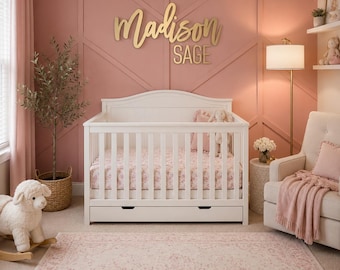 May include: A white wooden crib with a storage drawer sits against a dusty pink wall featuring geometric molding and the gold script text Madison SAGE. The crib includes a floral patterned mattress sheet and a plush toy.
