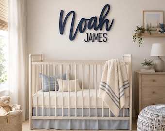 Puede incluir: Un letrero de pared de madera con el nombre Noah James en letras cursivas y de molde de color azul oscuro, montado sobre una cuna de madera clara. La cuna está vestida con una manta color crema con rayas azules, una almohada a cuadros azules.
