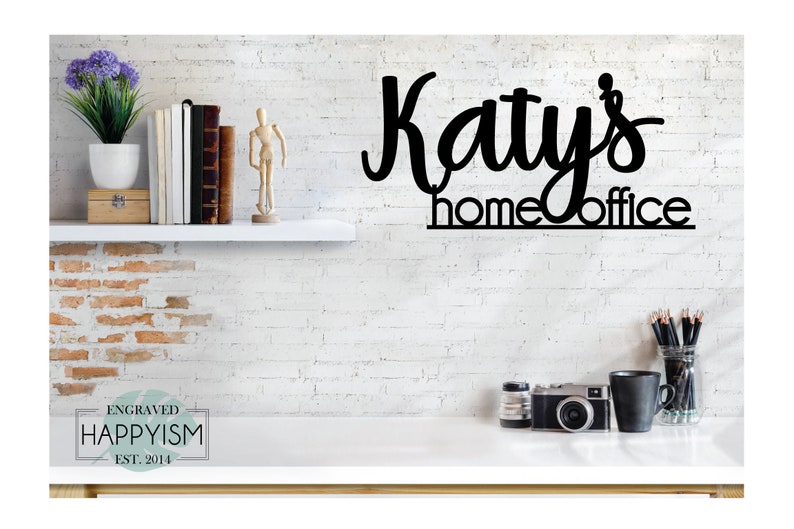 May include: Black metal sign with the text "Katy's home office" on a white brick wall background. The sign is hanging above a white desk with a camera, a mug, and a jar of pencils.