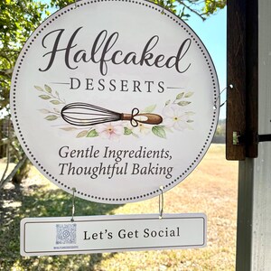 May include: A round sign for "Halfcaked Desserts" with a whisk and floral design. The sign also includes the text "Gentle Ingredients, Thoughtful Baking" and a smaller sign that says "Let's Get Social".