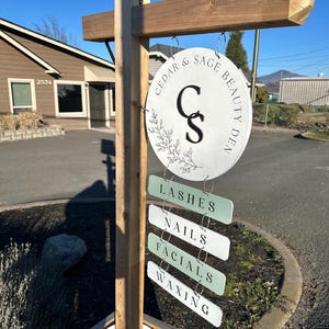 May include: A wooden signpost with a round white sign that reads "Cedar & Sage Beauty Den" with a floral design and the initials "C & S". Below are smaller signs that say "Lashes", "Nails", "Facials", and "Waxing".