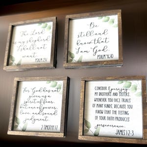 May include: Four wooden signs with inspirational bible verses and greenery. The signs feature the following verses: Psalm 23:1, Psalm 46:10, 2 Timothy 1:7, and James 1:2-3.