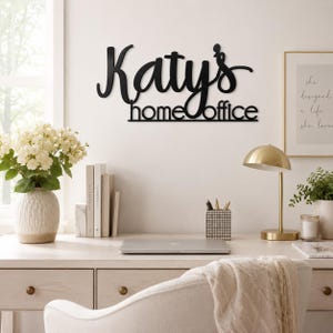 K&ouml;nnte beinhalten: Ein schwarzes Metall-Wandschild mit dem individuellen Schriftzug "Katy's home office" in einer Kombination aus Schreibschrift und serifenloser Typografie. Das Schild ist an einer Wand &uuml;ber einem Schreibtisch montiert und hat eine dunkle Oberfl&auml;che.
