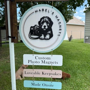 May include: A wooden sign for "Mabel's Magnets" featuring a black and white illustration of a dog with a camera. Below, three rectangular signs read "Custom Photo Magnets", "Loveable Keepsakes", and "Made Onsite". The sign is hanging outdoors.