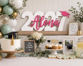May include: A graduation party display featuring large white wooden numbers 2027 with a pink script name Alona and a pink graduation cap topper. Includes a cake with a black cap, a letter board reading THE FUTURE IS BRIGHT, and cupcakes.