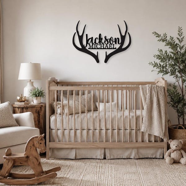 Pode incluir: Uma placa de parede preta com os nomes Jackson e Michael em letra cursiva, integrados num design em forma de duas hastes de veado simétricas. A placa está posicionada acima de um berço de madeira num quarto.