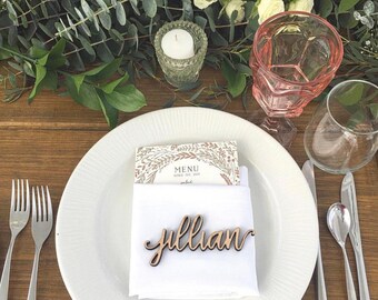 May include: A white dinner plate holds a folded white napkin topped with a wooden name tag reading Jillian. A menu card with floral illustrations sits behind the napkin. Silver forks, a knife, and a spoon are arranged on the table.