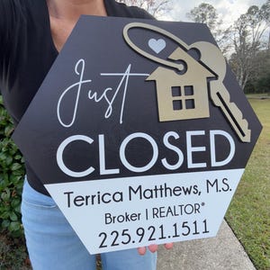 May include: A hexagonal sign with a black top and white bottom. It features the text Just CLOSED, a gold house icon with a key, and the name Terrica Matthews, M.S., Broker | REALTOR, and the phone number 225.921.1511 printed in black lettering.