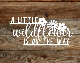 May include: A white wooden sign with the text "A little wildflower is on the way" with a floral design above the text.