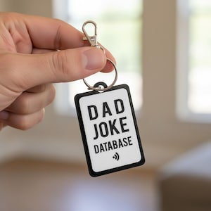 May include: A rectangular keychain with the words "DAD JOKE DATABASE" in black text on a white background. The keychain has a black border and a silver keyring and clasp.
