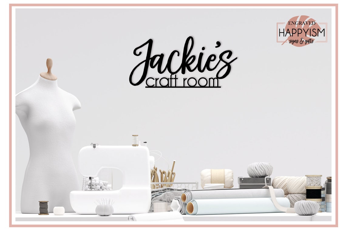 A custom craft room sign reads "Jackie's craft room" in script and non-script black text. The sign is hung on a white wall in a white room above a table covered in white craft accessories like a sewing machine, twine, yarn, vinyl, thread, and fabrics. There is a white sewing form on the right side.