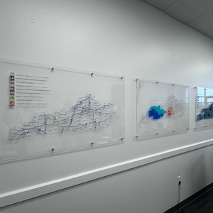May include: Three clear acrylic maps of Kentucky are mounted on a white wall. The leftmost map shows the state's highways and roads. The middle map shows the state divided into regions with different colored areas. The rightmost map shows a stylized design of the state.
