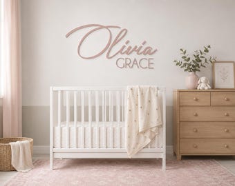 May include: A wall-mounted sign featuring the name Olivia Grace in a combination of cursive and block lettering. The sign is colored in a muted dusty rose and is positioned above a white crib with a patterned blanket draped over the side.