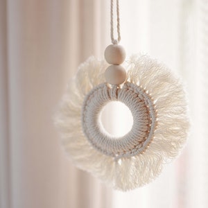 Könnte beinhalten: Cremefarbener Makramee-Wandbehang mit kreisförmigem Design. Die Mitte ist ein gewebter Ring, umgeben von einem Fransen aus geknoteten Fäden. Zwei Holzperlen sind über dem Kreis an einer passenden Schnur aufgehängt. Dekorative Wanddekoration.