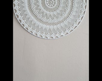 May include: A white, circular, crocheted wall hanging with a detailed, symmetrical design. The piece is mounted on a neutral-colored wall, creating a minimalist aesthetic. The intricate lace-like pattern adds texture and visual interest to the decor.