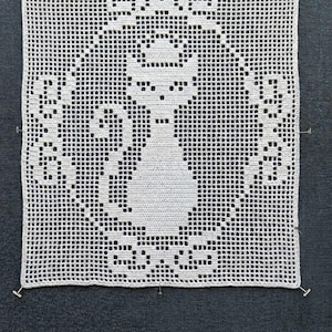 Puede incluir: Tapete cuadrado de ganchillo blanco con un diseño de gato. El gato está centrado, con un contorno detallado y elementos decorativos. El tapete está sujeto con alfileres a una superficie gris oscuro, mostrando el intrincado calado.