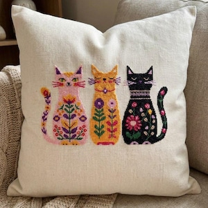 Puede incluir: Un cojín decorativo con fondo crema presenta tres gatos bordados. Los gatos son rosa, amarillo y negro, cada uno adornado con patrones florales en varios colores. El cojín está colocado sobre un sofá beige.
