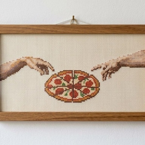 Puede incluir: Un marco rectangular de madera que contiene un bordado de punto de cruz sobre una tela clara. El diseño muestra dos manos extendiéndose hacia una pizza de pepperoni central, con las manos bordadas en tonos de hilo marrón y beige.