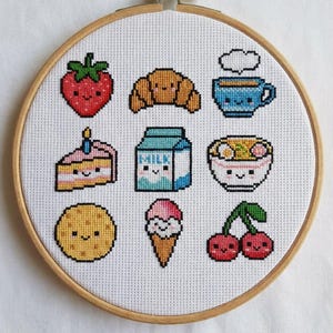 Puede incluir: Un bastidor de bordado de punto de cruz con nueve diseños lindos con temática de comida. Los diseños incluyen una fresa, un cruasán, una taza de café, un pastel, un cartón de leche, un cuenco de ramen, una galleta, un cono de helado y cerezas. El bordado está en un aro de madera.