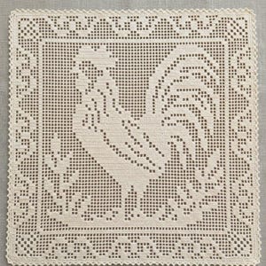Puede incluir: Un tapete de ganchillo cuadrado en color blanco roto que presenta un motivo central de gallo creado con una técnica de ganchillo de red. El diseño incluye una silueta de ave detallada rodeada por un borde geométrico con acabado festoneado.
