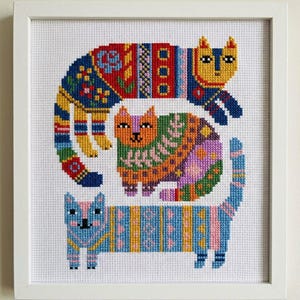 Puede incluir: Una obra de arte de punto de cruz enmarcada con tres gatos estilizados. Los gatos presentan patrones intrincados en colores vibrantes, incluyendo azul, amarillo, naranja y rosa. La obra está enmarcada en un marco blanco.