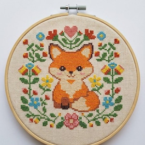 Puede incluir: Un bordado de punto de cruz de un zorro rodeado de flores y vegetación coloridas. El zorro es naranja y blanco, con una expresión dulce. El bordado está enmarcado en un aro de madera.