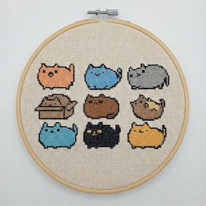 Puede incluir: Un bastidor de punto de cruz con nueve diseños de gatos pixelados en varios colores, incluyendo naranja, azul, gris, marrón y negro. Los gatos están dispuestos en un patrón de cuadrícula sobre una tela beige, enmarcada por un aro de madera.