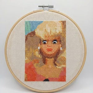 Puede incluir: Un retrato de punto de cruz de una mujer con cabello rubio, labios rojos y ojos azules, enmarcado en un bastidor de bordado de madera. La obra presenta una parte superior roja y un fondo colorido, mostrando una estética retro.