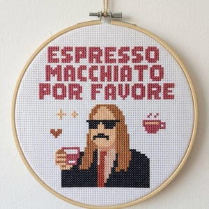 Puede incluir: Arte de punto de cruz en un aro de madera. Muestra una figura pixelada con gafas de sol y bigote, sosteniendo una taza de café. El texto "ESPRESSO MACCHIATO POR FAVORE" está bordado en rojo. Incluye corazones y estrellas.