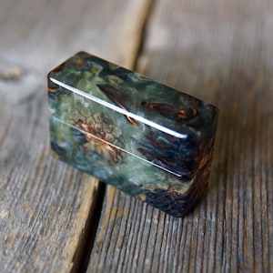 Peut inclure: Un étui à briquet rectangulaire doté d'une finition polie avec un motif marbré dans des tons de vert forêt profond, de brun foncé et des touches d'ambre. La surface présente une texture organique complexe avec des inclusions sombres.