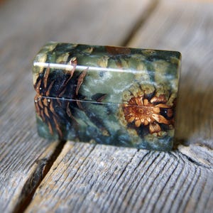 May include: A rectangular box with a smooth, polished finish featuring a dark green and blue resin body embedded with natural brown pine cone segments. The lid and base are aligned, showcasing a seamless transition between the organic textures.